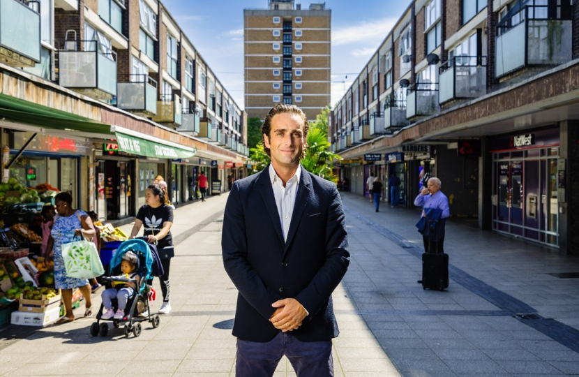 Alex Clarkson, smartly dressed, stood in Stevenage Town Centre. He is flanked by retails shops on either side. Directly behind him is a large town block of flats.