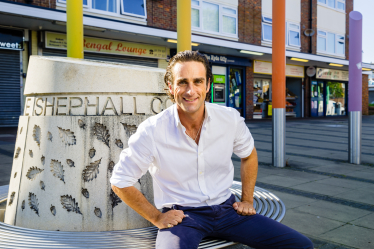 Alex sat at the Hyde Shops. Behind him is a concrete sculpture engraved with 'Shehall' as well as several different coloured pencil statutes