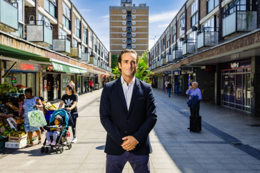 Alex Clarkson, smartly dressed, stood in Stevenage Town Centre. He is flanked by retails shops on either side. Directly behind him is a large town block of flats.